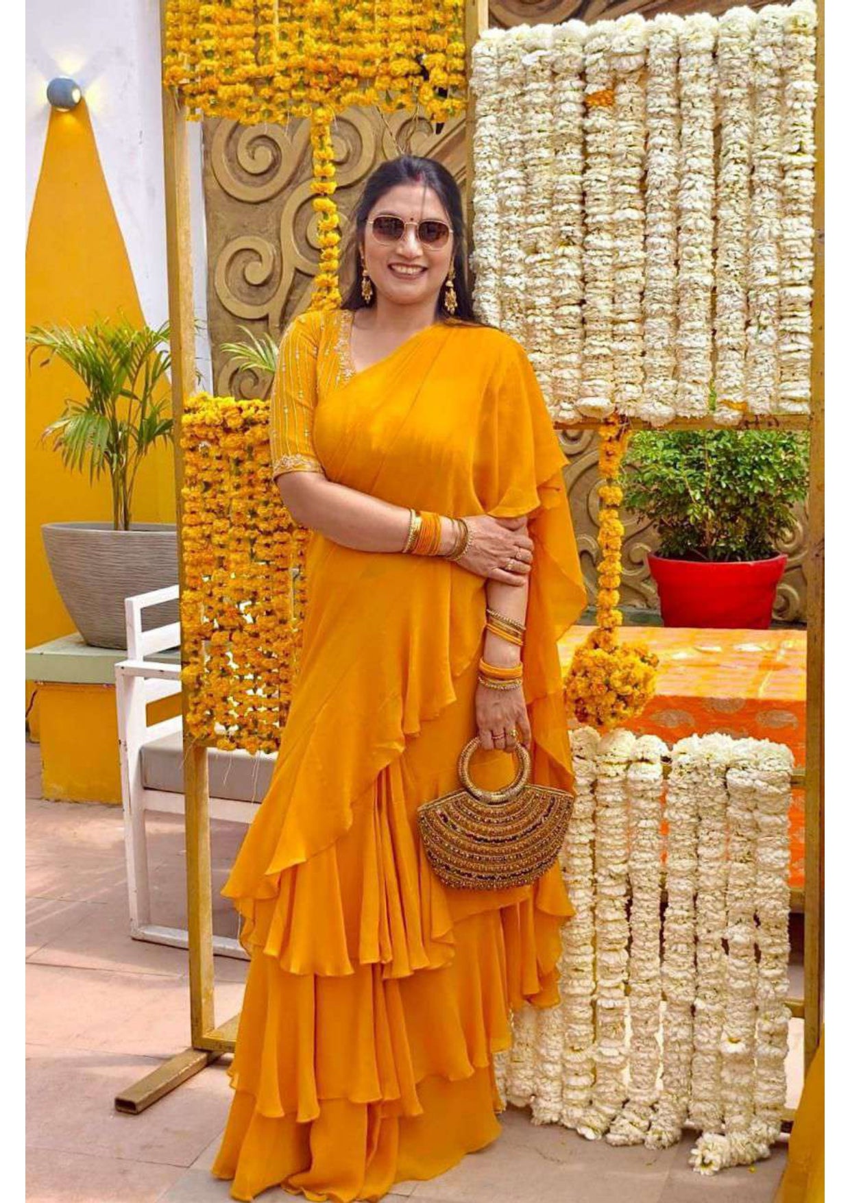 Woman in yellow saree poses at a marigold-themed event with floral decorations and a woven handbag.